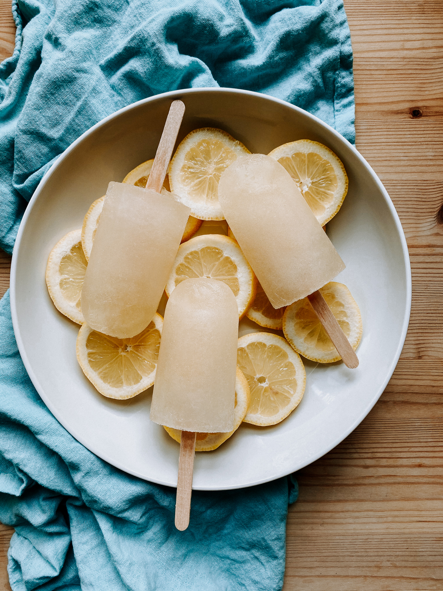 a white bowl with slices of lemon and lemon ic epops resting on a blue towel on a wooden surface