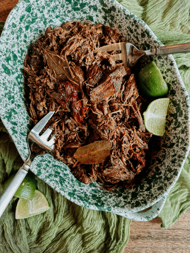 Shredded barbacoa beef served in a green speckled dish with bay leaves, chipotle peppers, and lime wedges.