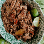 Shredded barbacoa beef with bay leaves and limes in a green speckled serving dish.