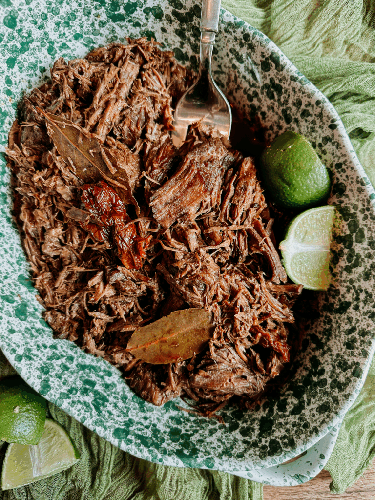 Shredded barbacoa beef with bay leaves and limes in a green speckled serving dish.