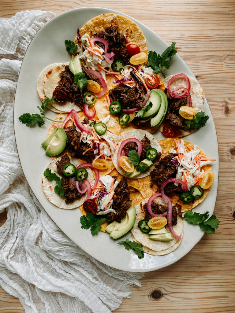 A platter of barbacoa beef tacos topped with avocado, slaw, pickled onions, and fresh cilantro.