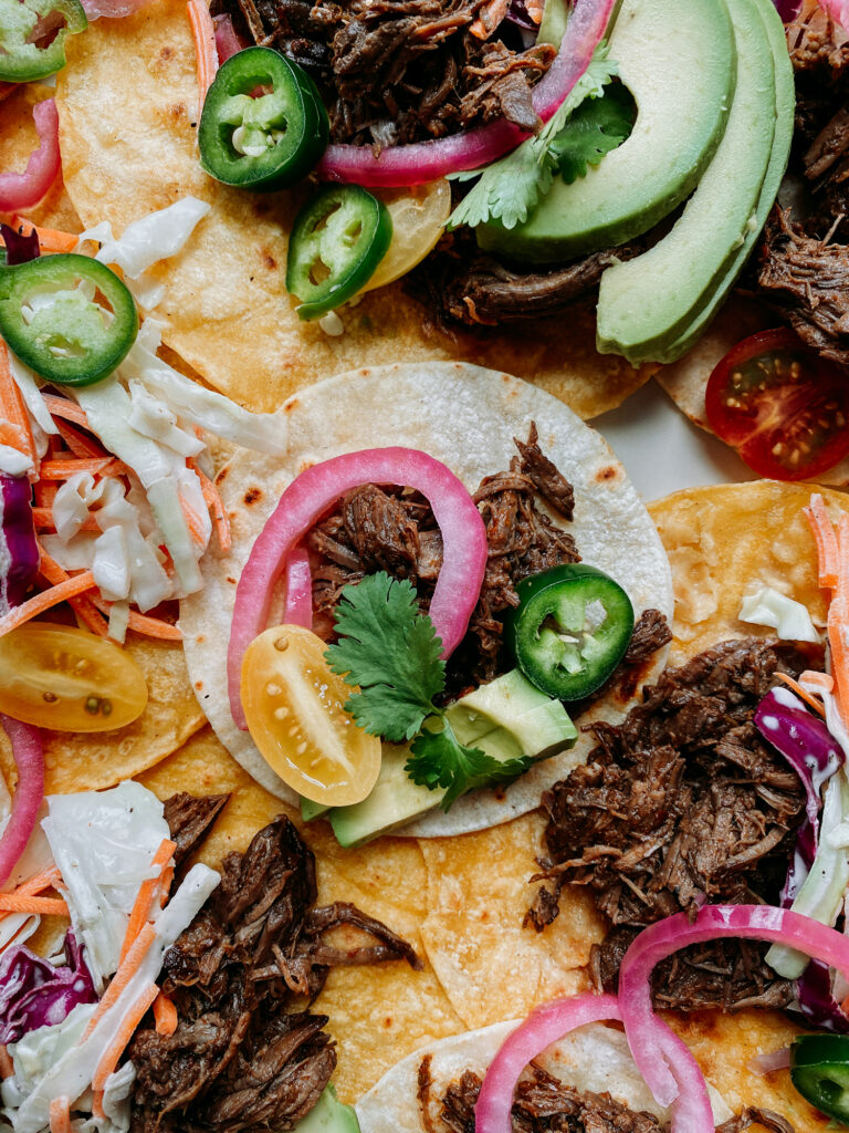 Overhead shot of barbacoa tacos with avocado slices, jalapeños, slaw, and pickled onions on toasted tortillas.