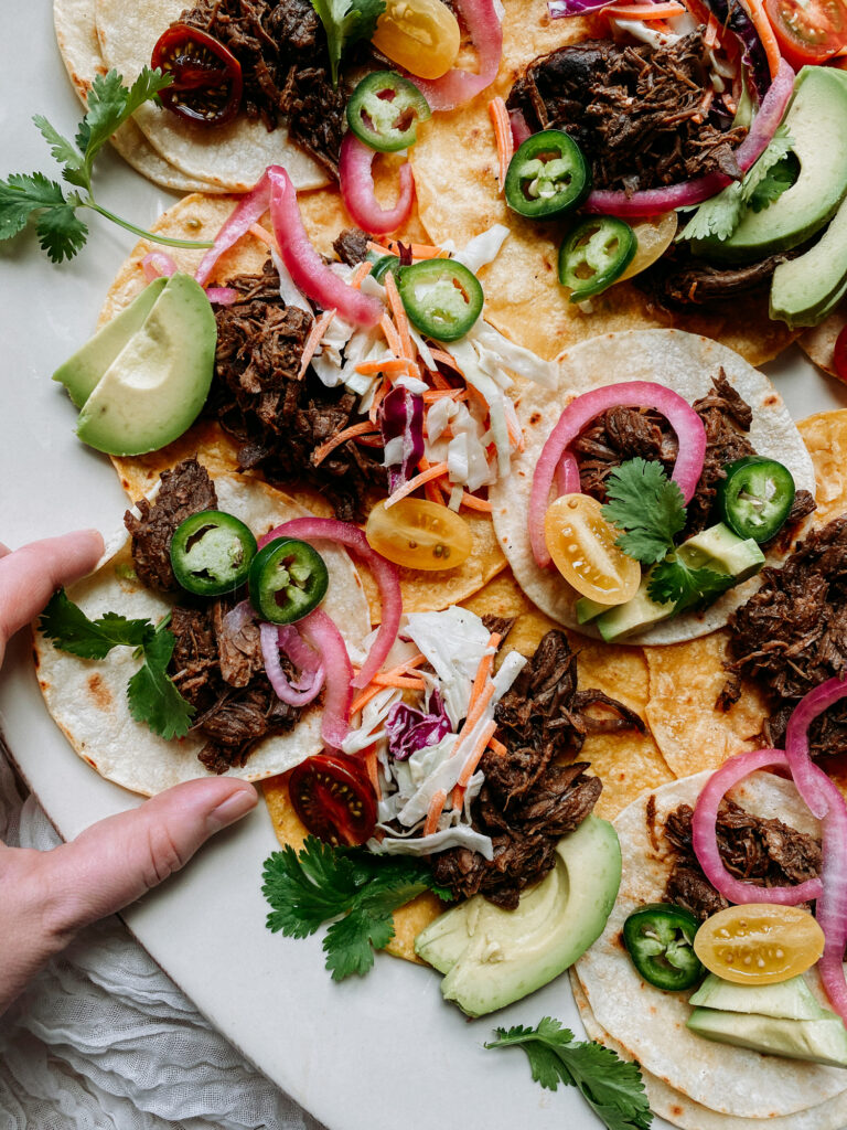 Close-up of barbacoa tacos with colorful toppings including jalapeños, avocado, and cherry tomatoes.