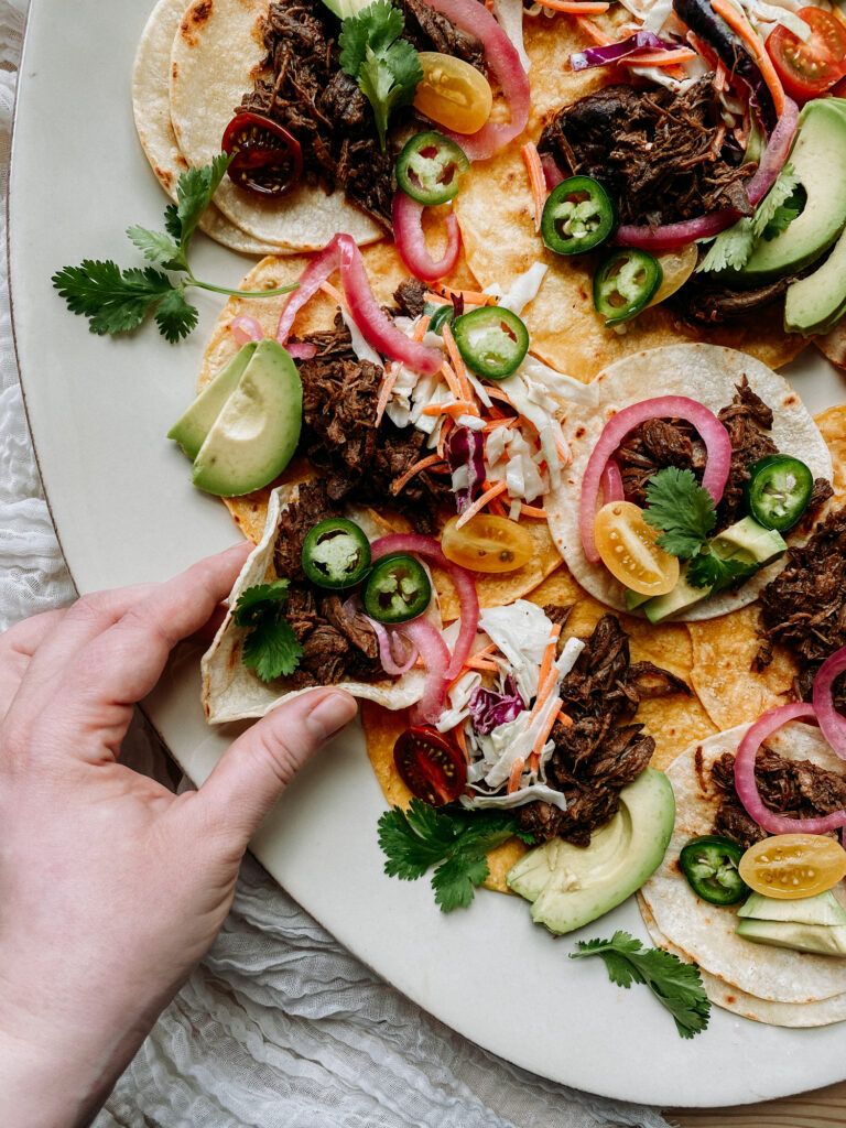 Hand reaching for a barbacoa taco from a serving platter filled with colorful, garnished tacos.