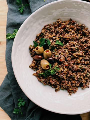 several whole green and kalamata olives resting on top of a pile of processed olives in a white bowl