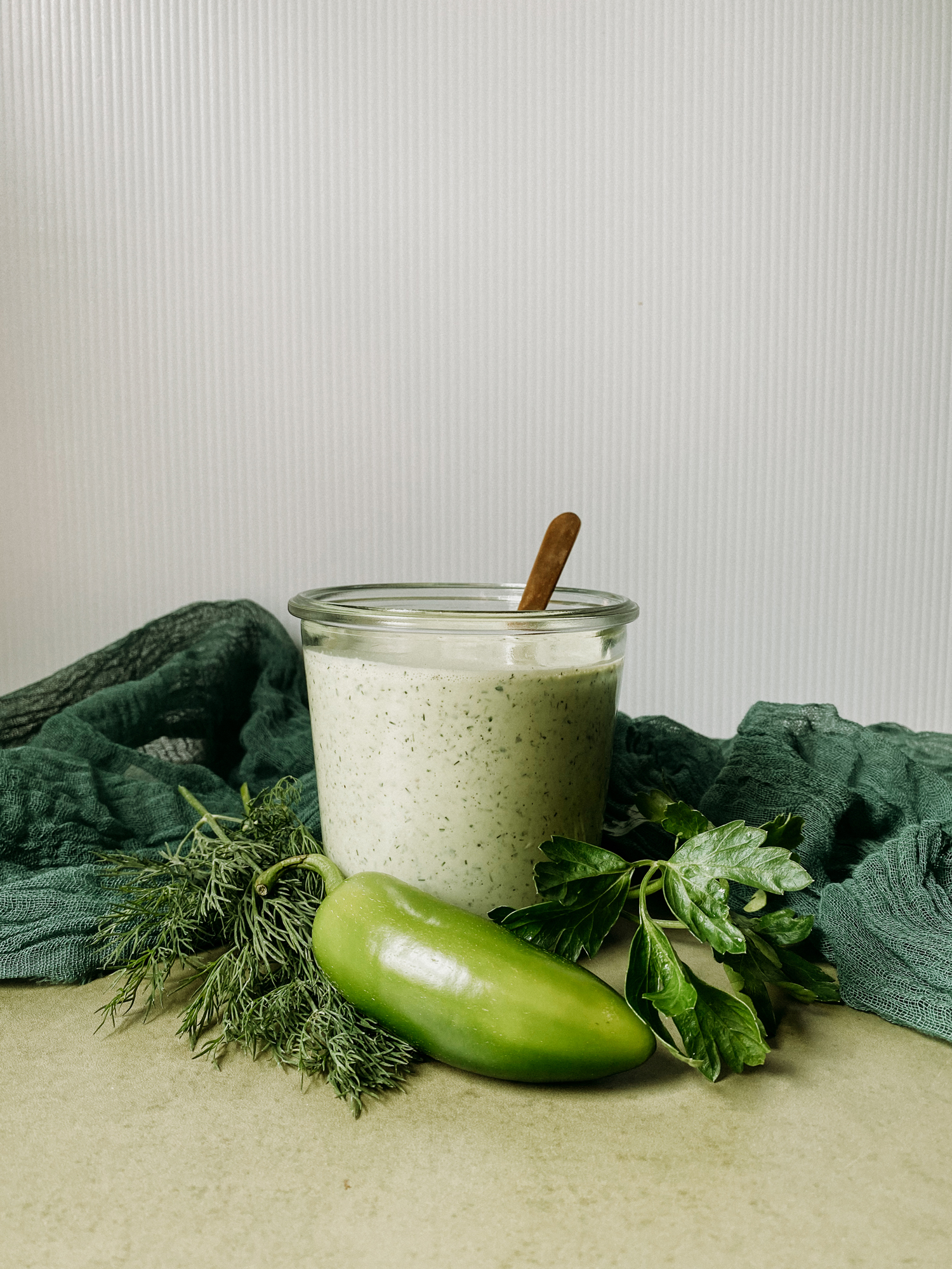a side view of a glass container of Buttermilk Jalapeno Ranch Dressing with jalapenos and herbs surrounding the container