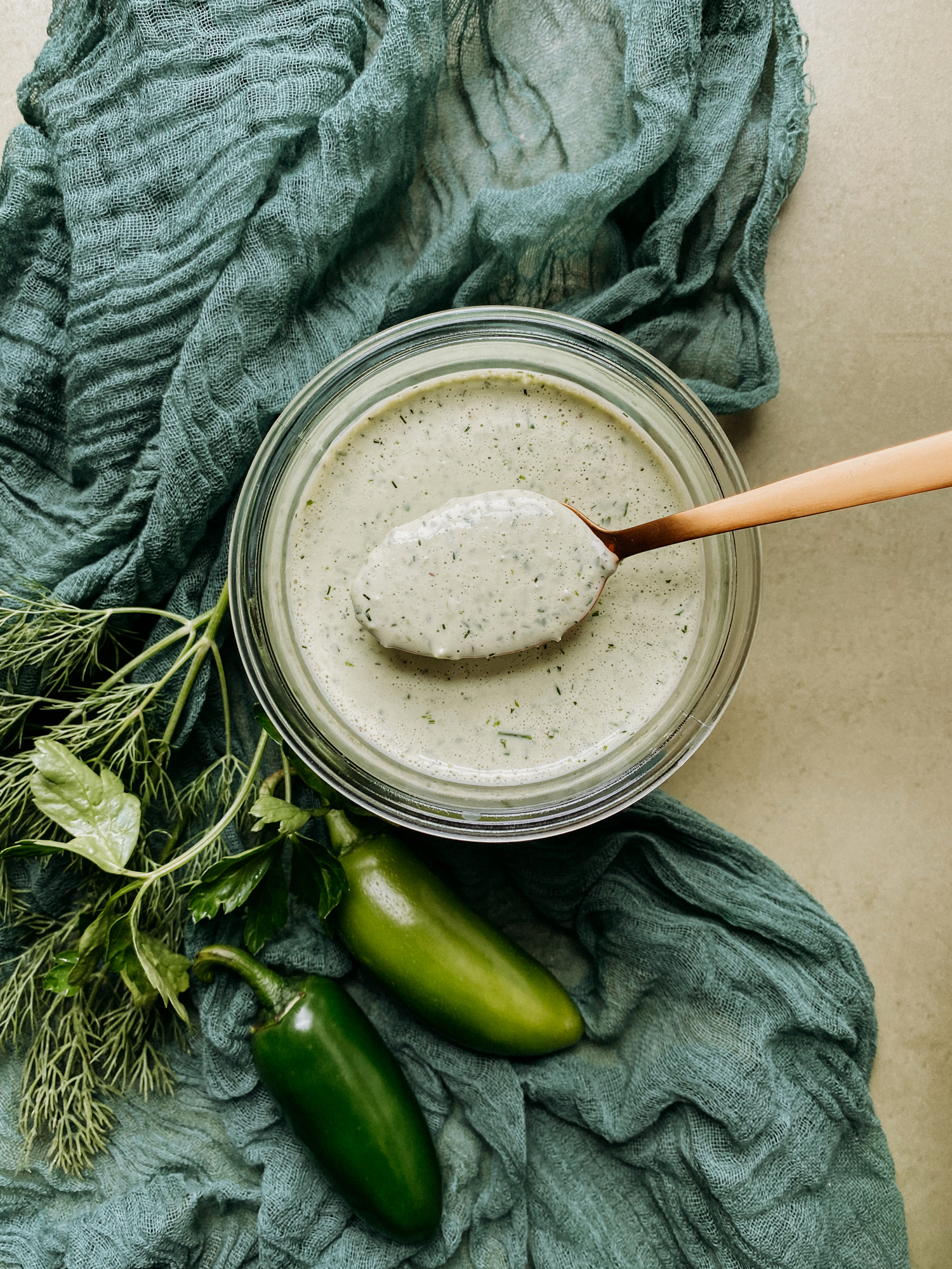 a gold spoon holding a spoonful of Buttermilk Jalapeno Ranch Dressing over the top of a glass container with a green cloth underneath