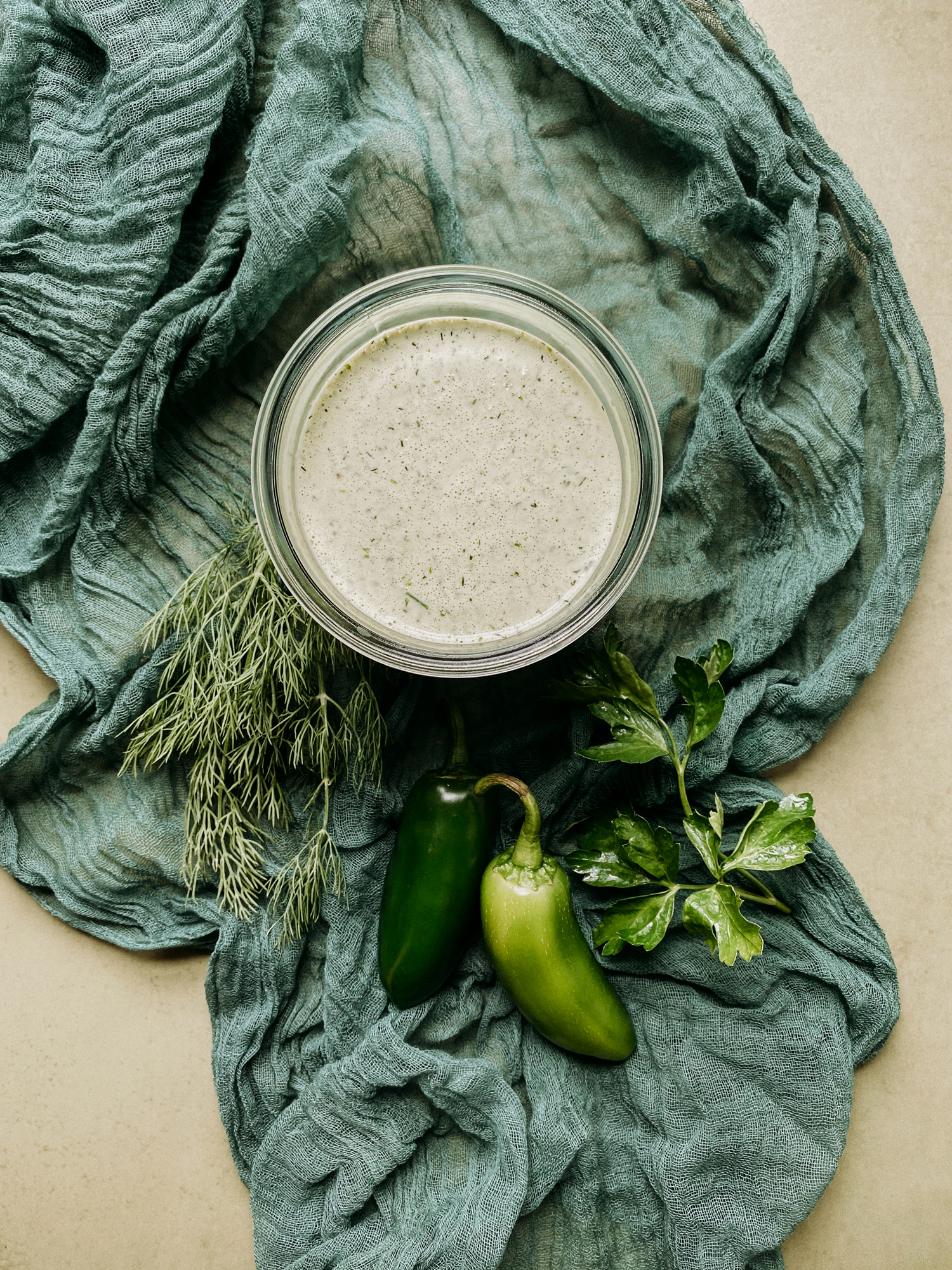 a green cloth with a glass container of Buttermilk Jalapeno Ranch Dressing resting on top alongside fresh jalapenos and herbs