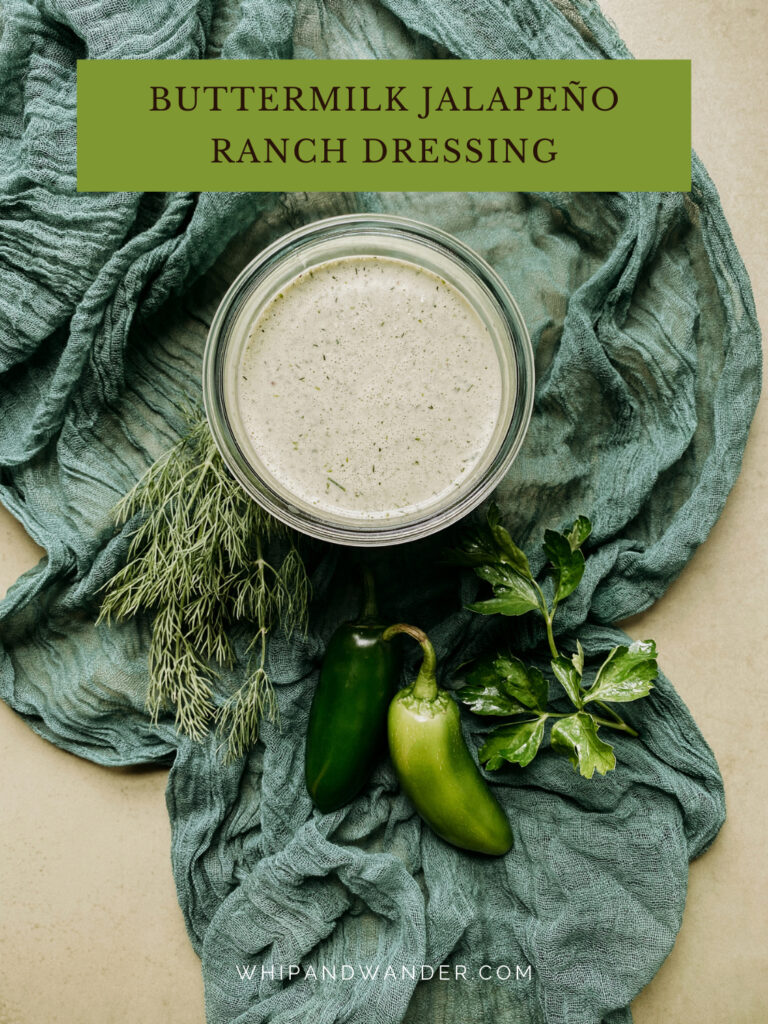 a glass jar of Buttermilk Jalapeno Ranch Dressing resting on a green cloth with herbs and fresh jalapenos