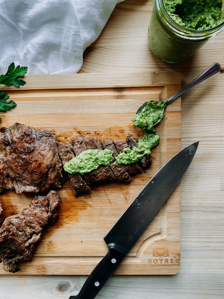 Grilled Skirt Steak slices with avocado chimichurri sauce on top on a wooden cutting boar with a spoonful of sauce and a knife resting nearby