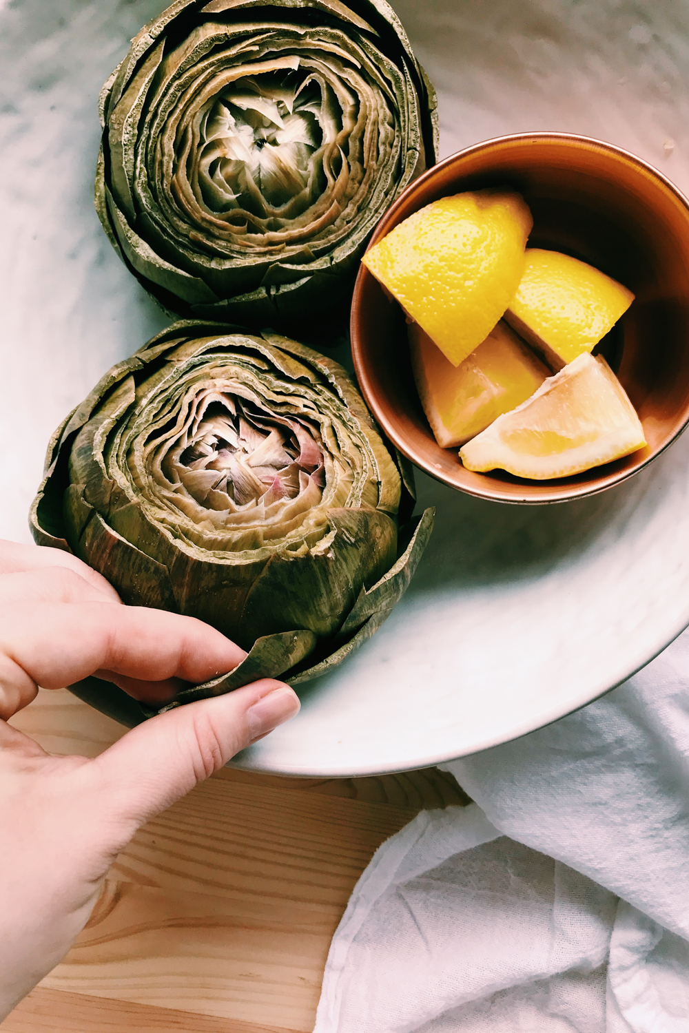 a white bowl with instant pot artichokes and a hand pulling leaves off with lemons int he bowl