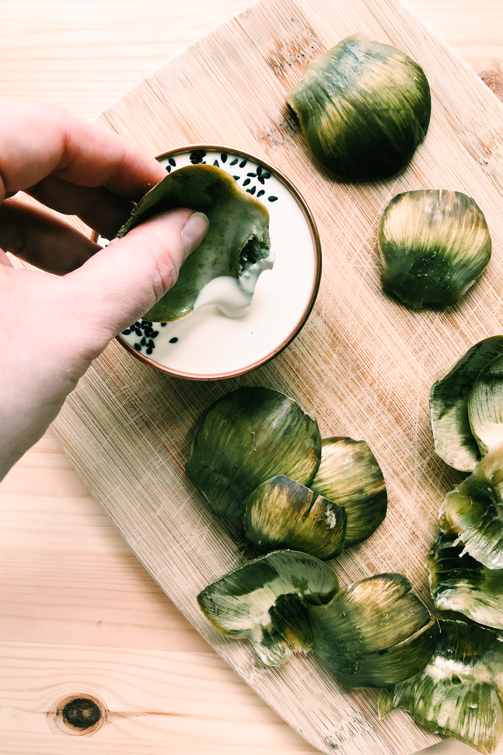 a hand dipping an artichoke leaf in a bowl of mayo vinegar dipping sauce