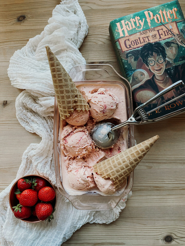 a harry potter book sitting next to a glass dish of strawberry buttermilk ice cream with a metal ice cream scoop and two cones resting in the dish. a bowl of strawberries resting nearby
