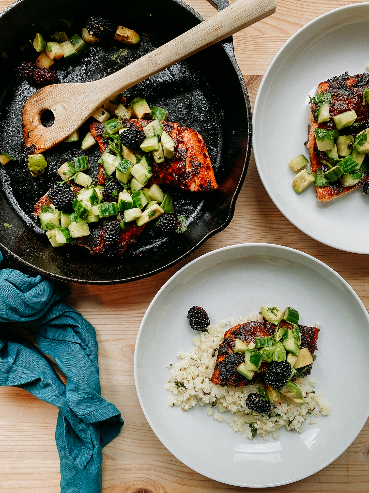 a cast iron pan with salmon and green citrus avocado berry salsa resting next to two white bowls that have salmon, salsa, and cauliflower rice