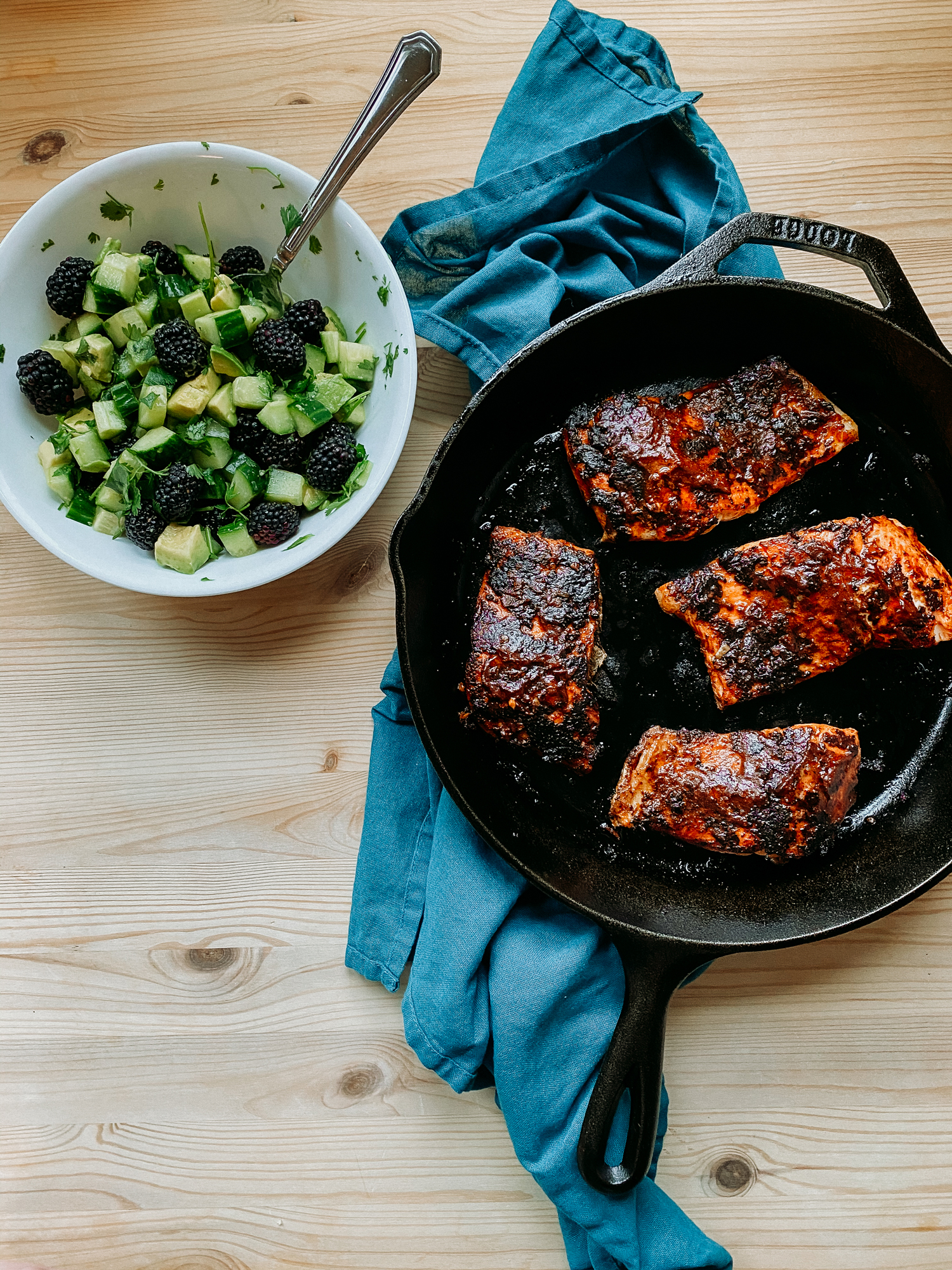 a cast iron pan resting on a blue towel on a wooden table that has spicy salmon adodo and a white bowl with blackberry salsa sitting nearby