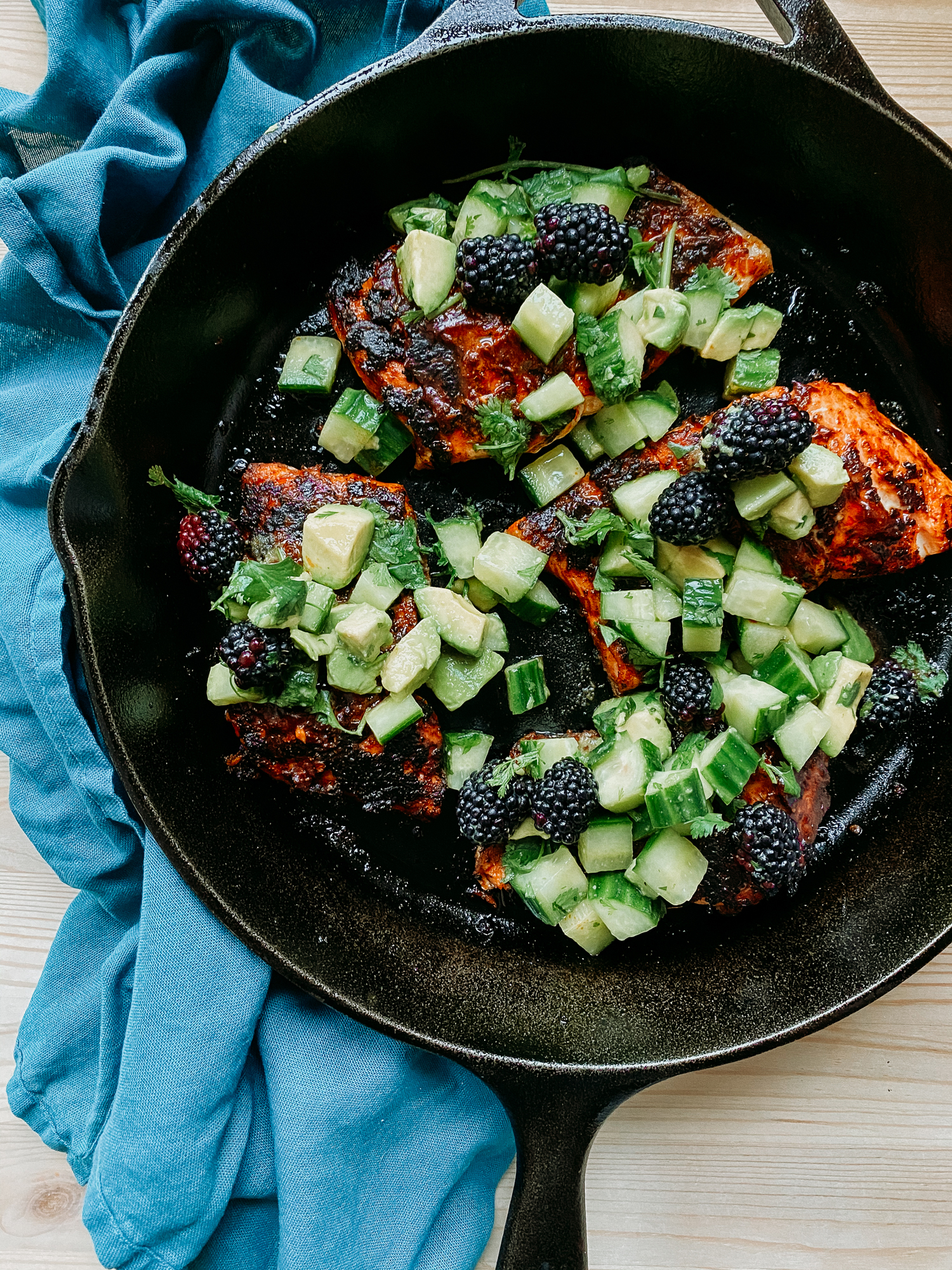 a black cast iron pan with salmon with adobo sauce and blackberry cucumber avocado salsa on top resting on a blue towel
