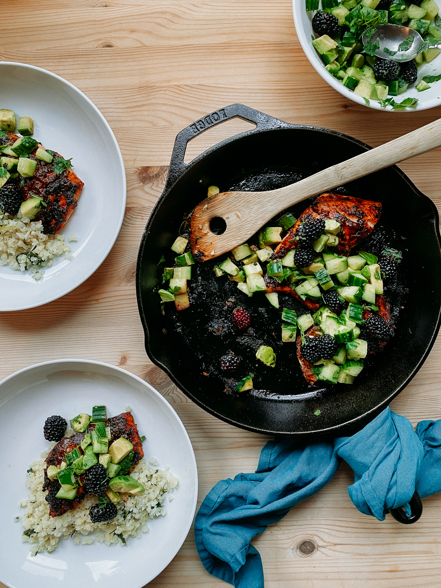 a cast iron pan with a blue towel wrapped around the handle that holds salmon fillets with adobo sauce and salsa that has cucumbers, avocados, and blackberries, a wooden serving utensil rests int eh pan and two white bowls rest to the left with salmon in them