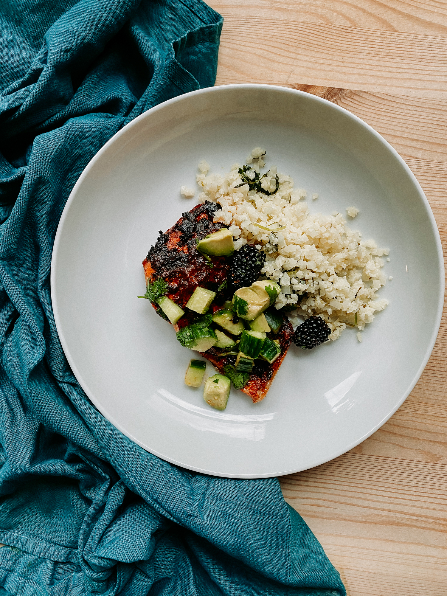 a filet of salmon with adobo sauce topped with blackberry, lime, cucumber, avocado salsa resting on cauliflower rice in a white bowl on top of a wooden surface with a blue towel