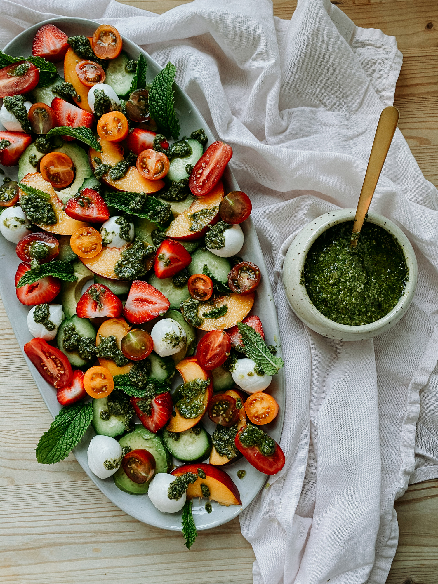 strawberry peach summer salad with cucumbers and peso on a white serving dish with a dish of pesto and a gold spoon resting on a white towel