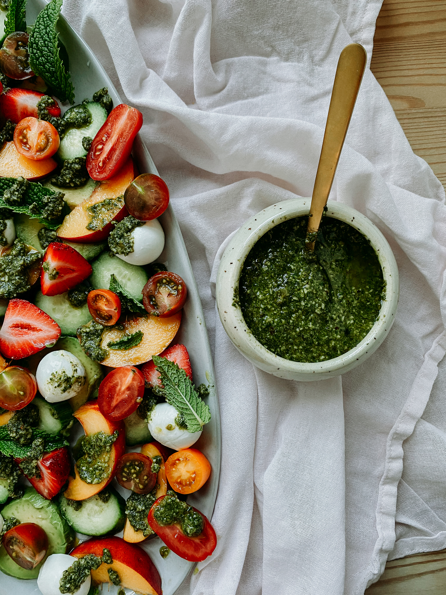 basil walnut pesto in a dish witha gold spoon sitting next to a platter of summer salad with strawberries and peaches