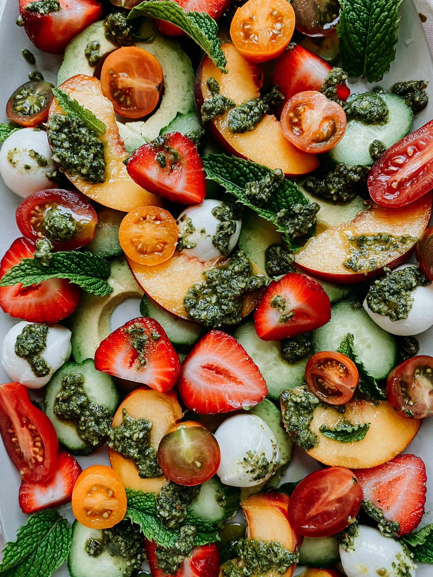 a strawberry and peach salad with cucumbers, tomatoes, avocado, mozzarella, and pesto on a white surface