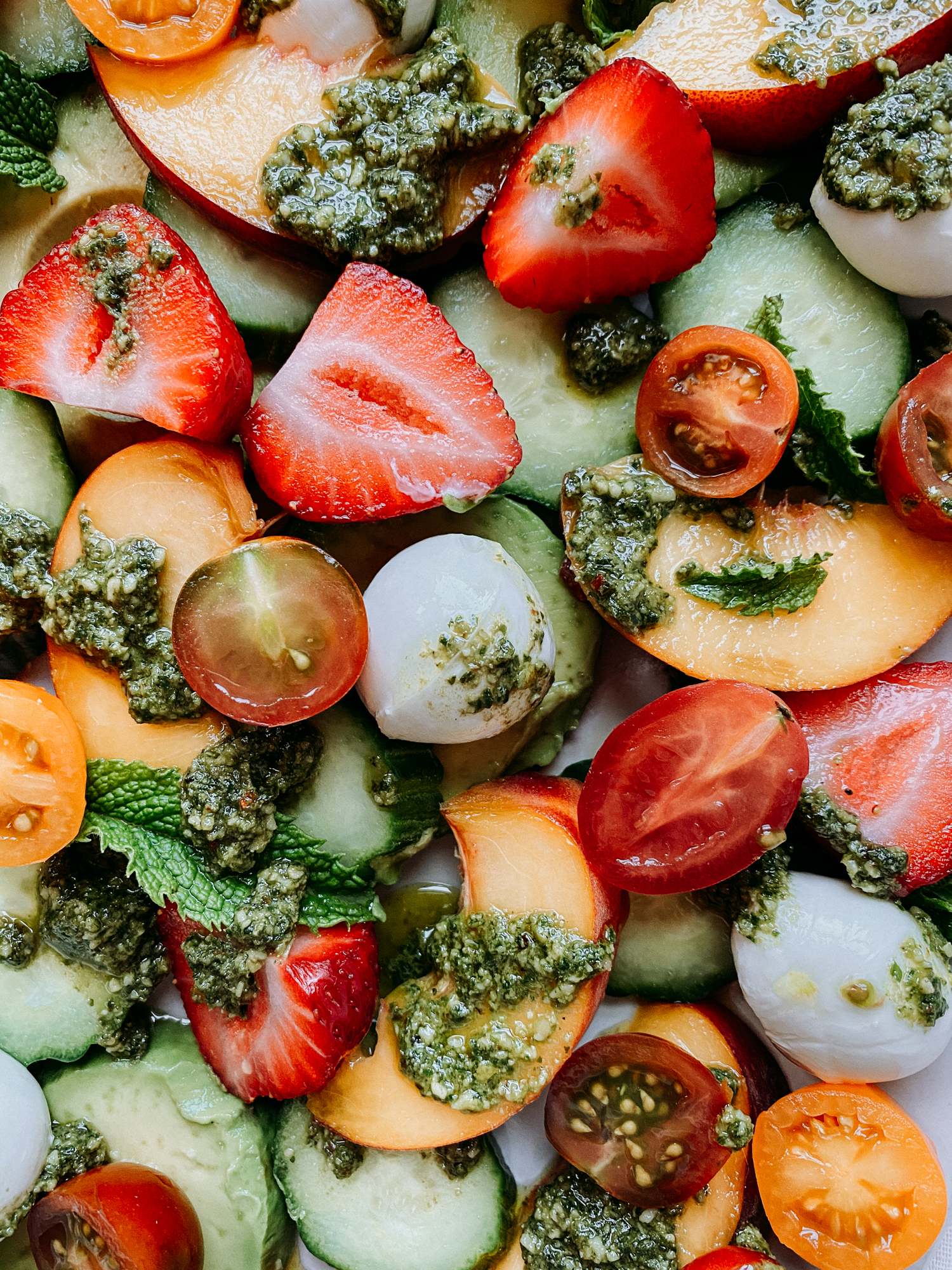 closeup of mozzarella balls, sliced strawberryies, sliced peaches, cucumbers, avocado slices, sliced tomatoes, and pesto