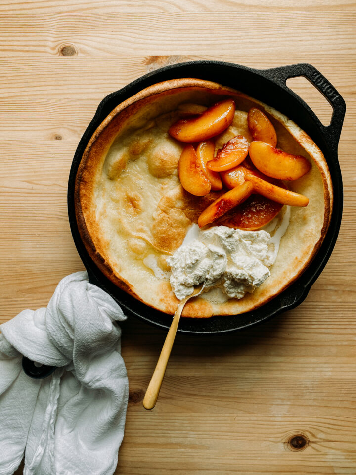 Peaches and fresh whipped cream on top of a Buttermilk Dutch Baby in a cast iron skillet resting on a wooden surface