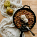 a spoon resting in a cast iron pan containing Spiced Pear Crisp and scoops of vanilla bean ice cream resting on a white towel with two fresh pears