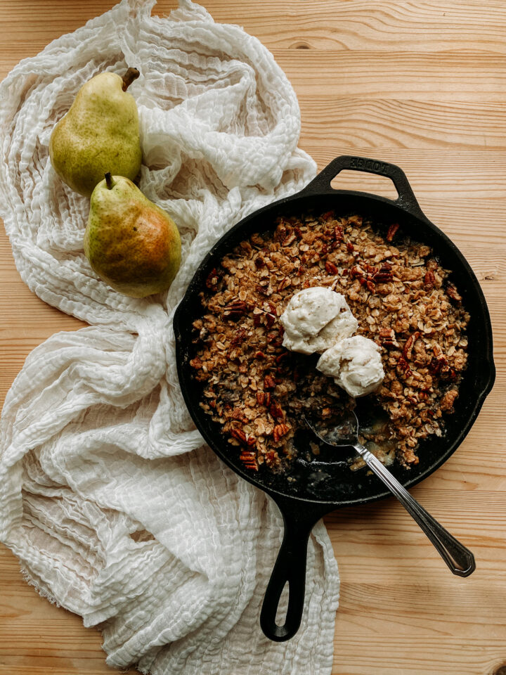 a spoon resting in a cast iron pan containing Spiced Pear Crisp and scoops of vanilla bean ice cream resting on a white towel with two fresh pears