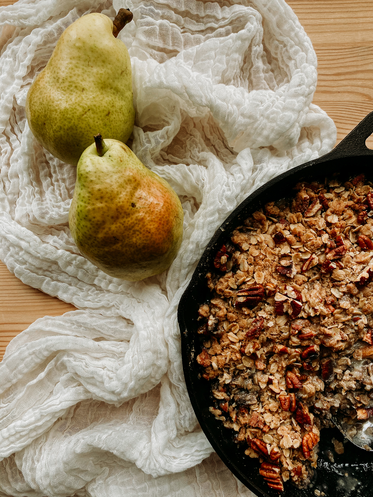 two pears resting on a neutral colored towel next to a cast iron pan of Spiced Pear Crisp