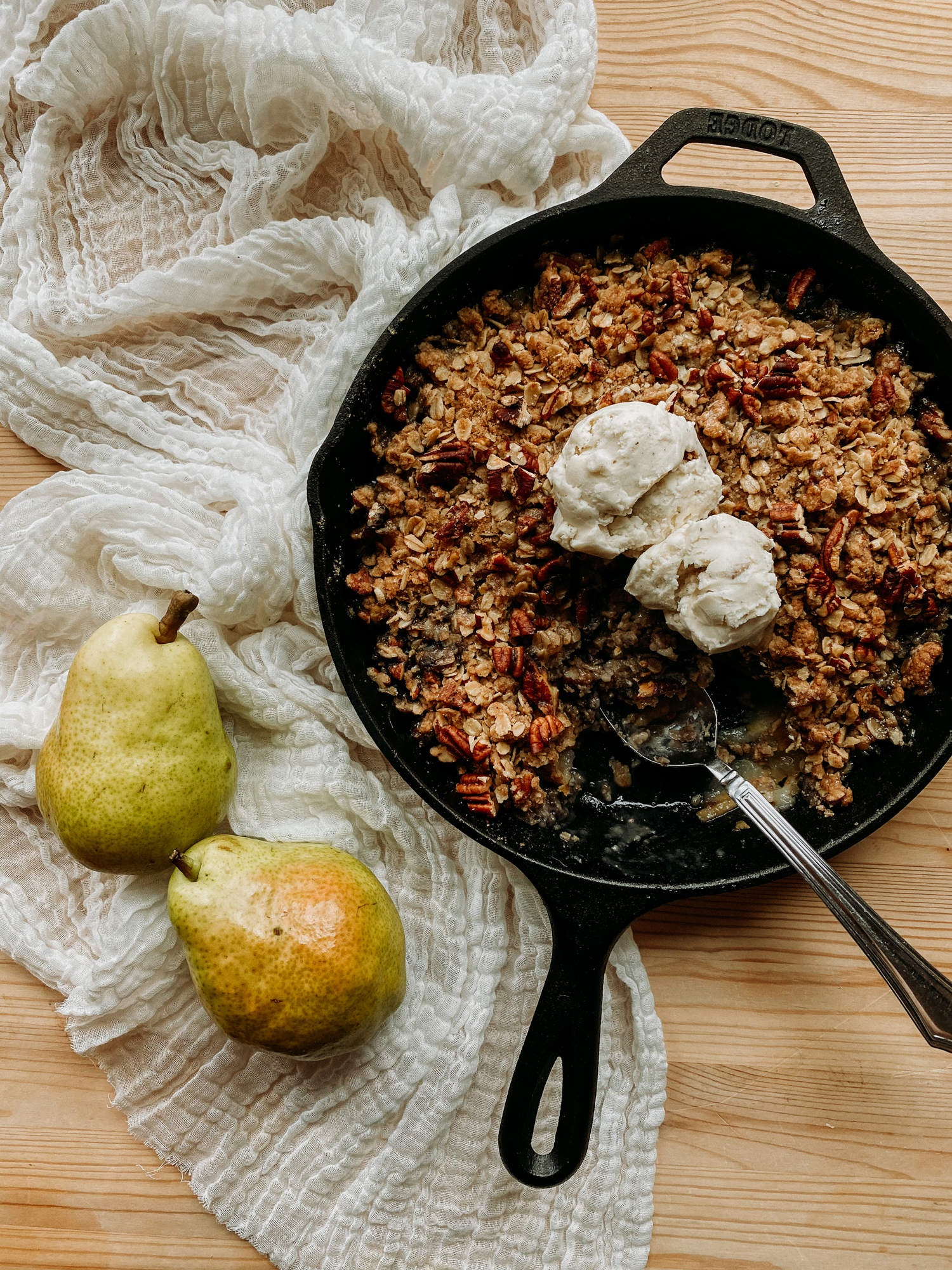 spiced pear crisp with ice cream in a cast iron pan resting next to two pears on a neutral colroed towel