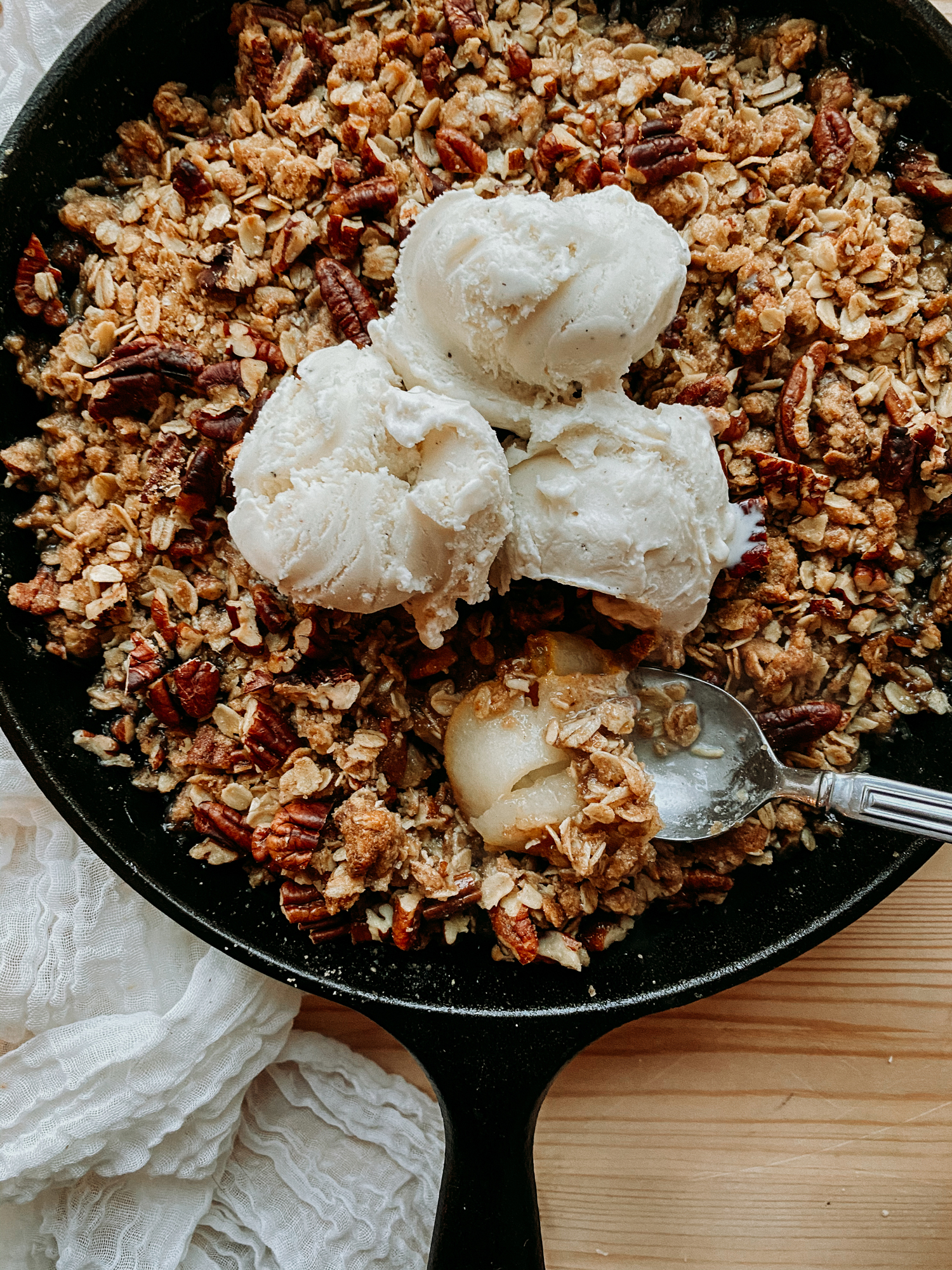 a spoon resting in a cast iron pan of Spiced Pear Crisp that contains three scoops of vanilla ice cream