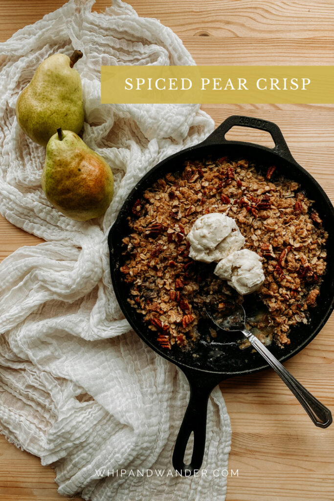 a cast iron pan of Spiced Pear Crisp resting next to a iece of cream colored cloth with two fresh barlett pears resting on top