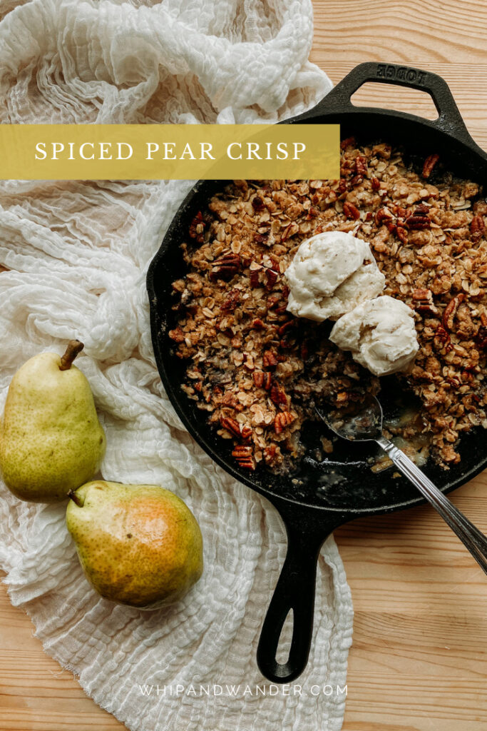 a vanilla ice cream topped pear crisp with spices in a cast iron pan with a spoon resting in the pan and two fresh pears resting next to the dish on a wooden table with a piece of cream colored cloth