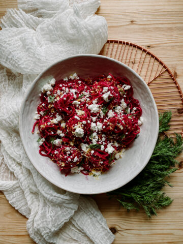 a large white bowl with bright pink pasta, goat cheese, and dill inside, resting on top of a white towel on a copper tray with a large swatch of fresh dill weed on a wooden surface
