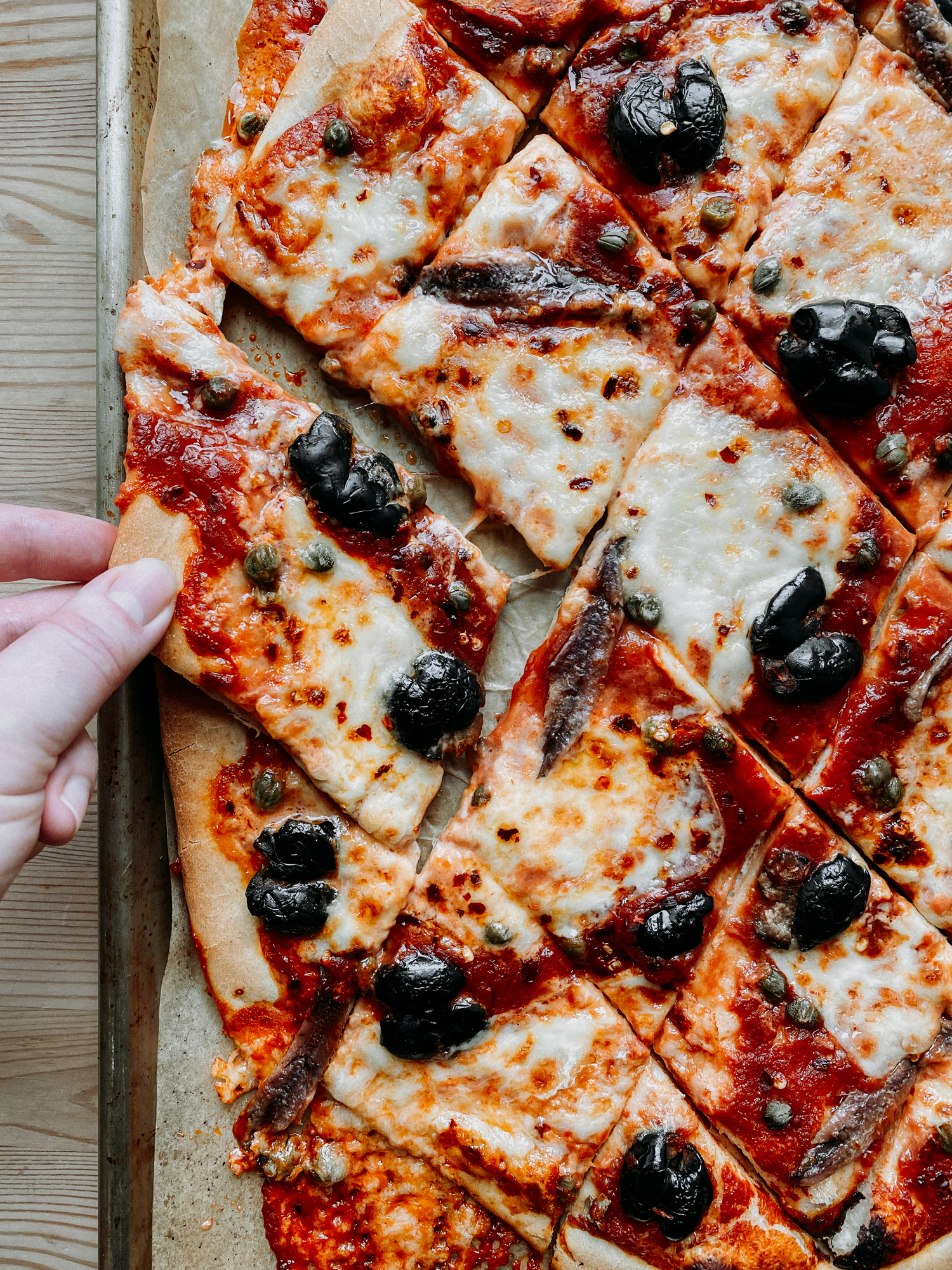 a hand pulling a slice of pizza off of a baking sheet