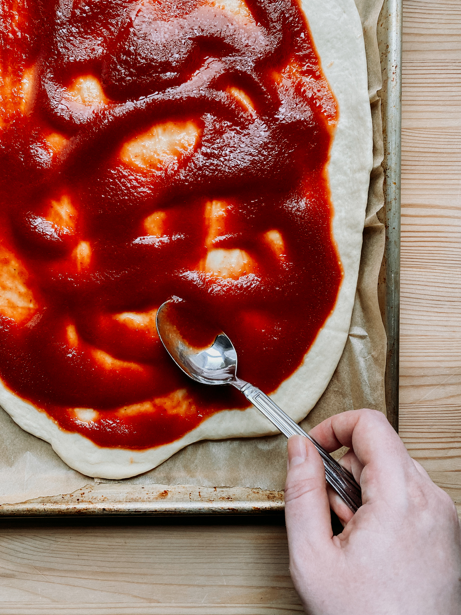 a hand using a spoon to spread tomato sauce over a pizza crust