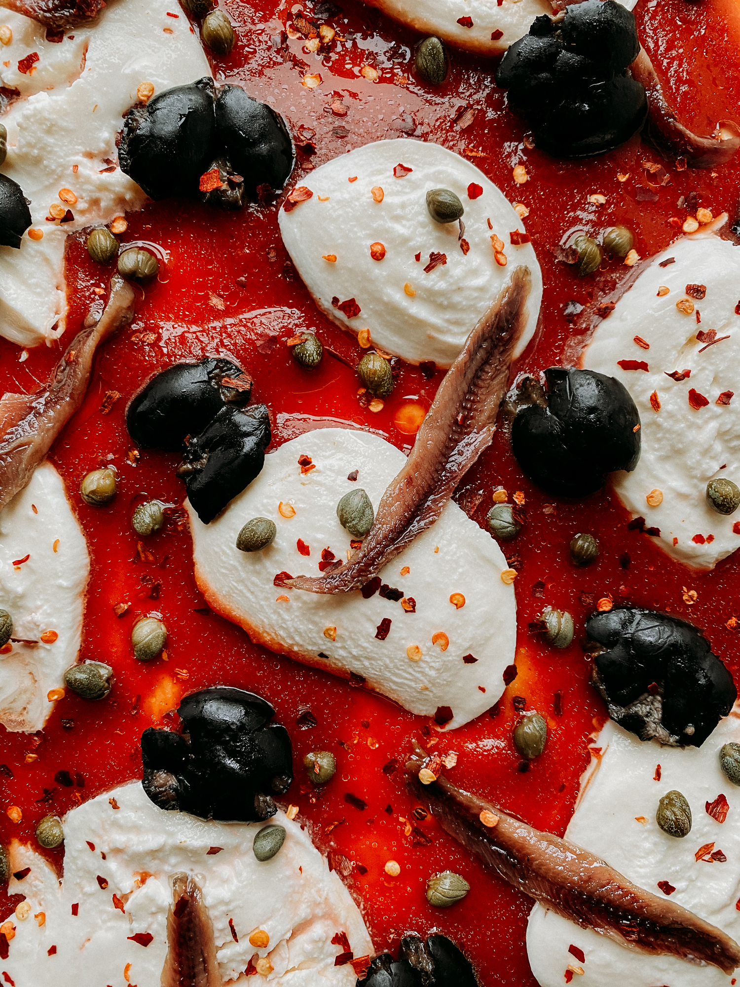 closeup of anchovies, olives, capers, and mozzarella cheese on a red sauce topped pizza crust