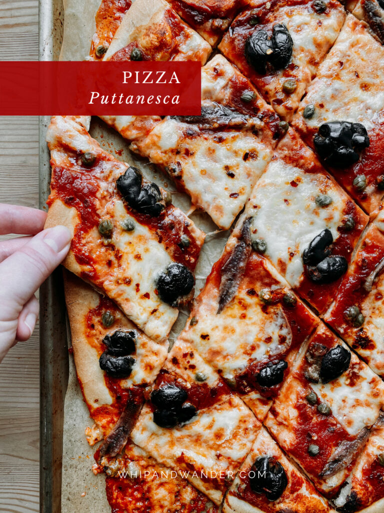 a hand pulling a slice of Pizza Puttanesca off of a parchment lines baking sheet