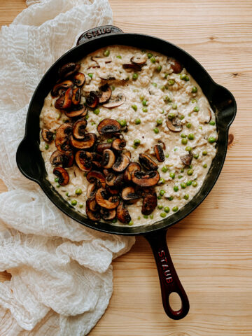 a dark red staub skillet filled with Risotto with Mushrooms and Peas resting on a gauzy fabric topped wooden surface