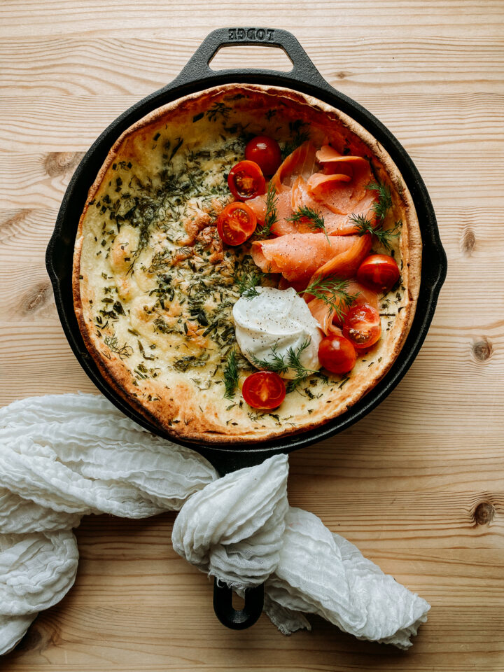 A Savory Buttermilk Herb Dutch Baby in a cast iron skillet topped with smoked salmon, tomato, and mascarpone resting on a wooden surface