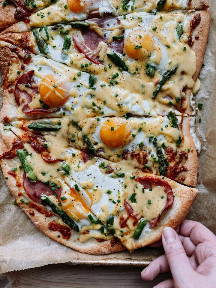 a hand grabbing a slice of Eggs Benedict Pizza off of a baking tray