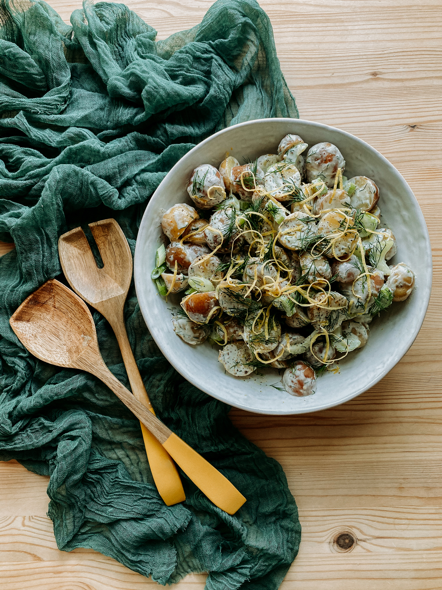 a large bowl filled with Creamy Dijon Dill Potato Salad next to two yellow tipped serving utensils resting on a green cloth