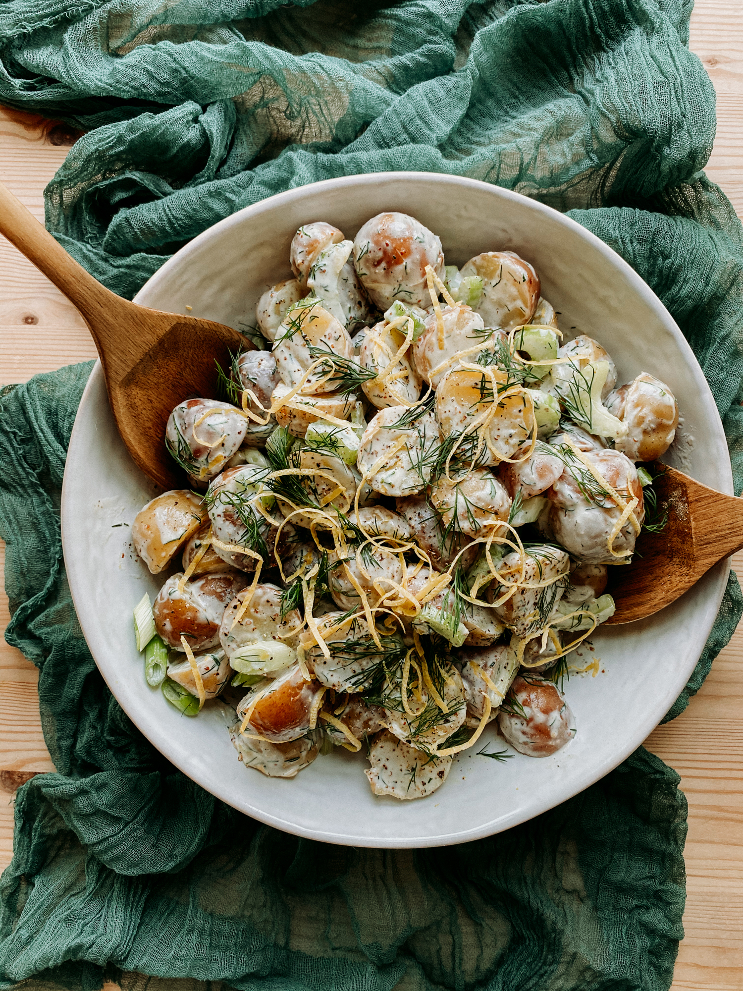 a white dish filled with potato salad and wooden serving utensils resting on a green piece of fabric