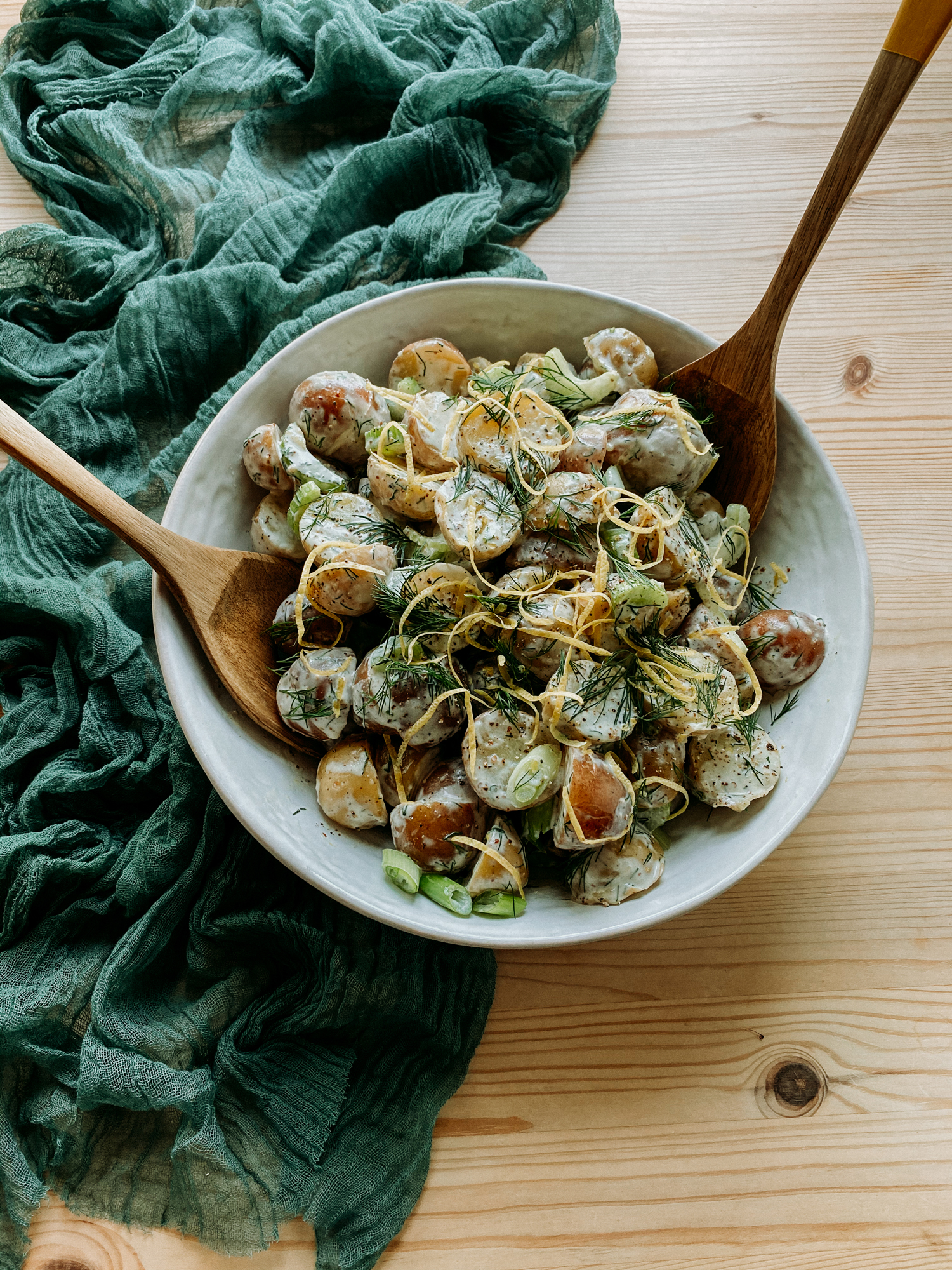 wooden serving utensils resting in a lareg white bowl of potato salad on a green cloth lined table