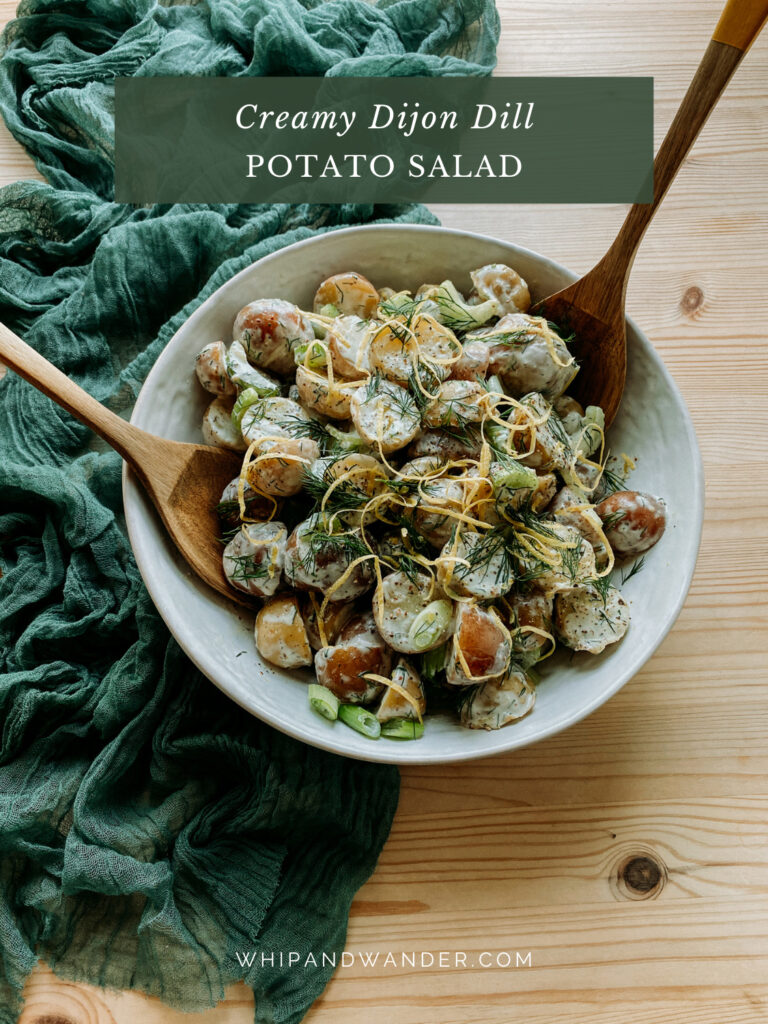 two wooden serving utensils resting in a white bowl filled with Creamy Dijon Dill Potato Salad
