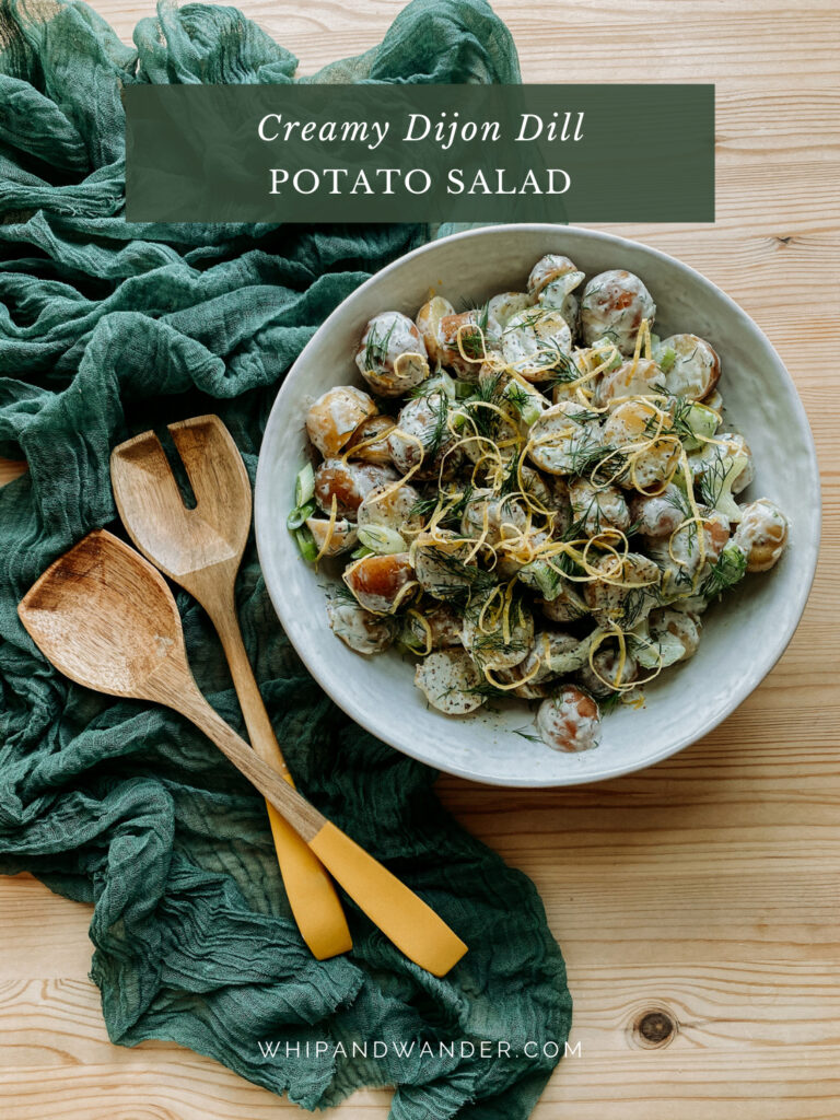 Creamy Dijon Dill Potato Salad in a large white serving bowl resting on a green cloth with yellow tipped wooden salad servers resting nearby