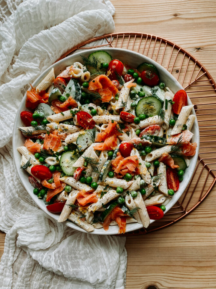 Penne pasta with tomatoes, cucumbers, smoked salmon, and herbs in a white bowl on a brass tray with a white cloth
