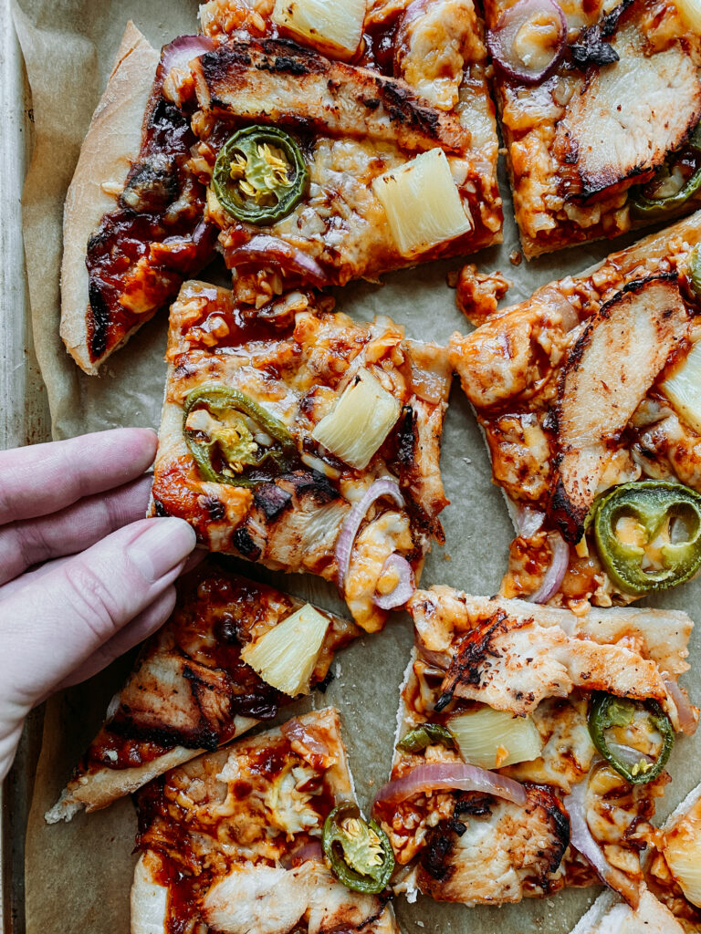 a hand grabbing a slice of BBQ Chicken Pizza from a baking sheet