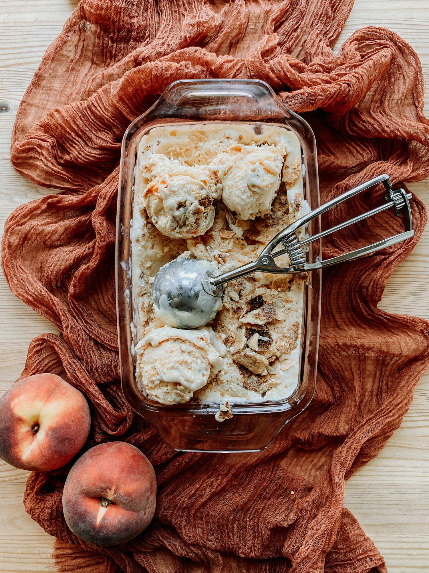 Buttermilk Peach Pie Ice Cream in a pink glass loaf pan resting on a dark peach colored cloth with a metal ice cream scoop in the pan